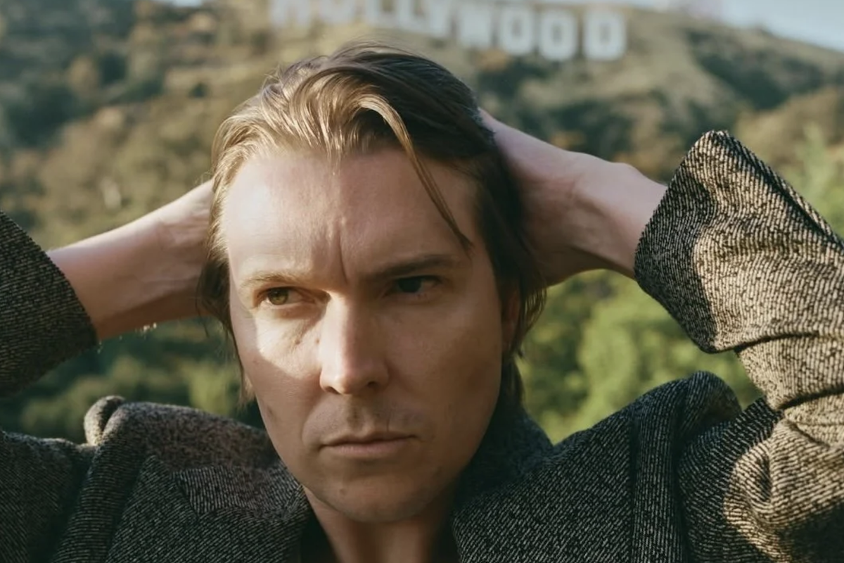 Musician Alex Cameron posing outdoors with his hands behind his head and the Hollywood Sign visible in the background.